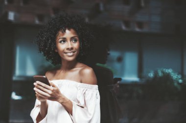 Neşeli bir kıvırcık saçlı Genç Brezilyalı kadın bir ayna duvara yaslanmış ve kopya alanı bölgesi metniniz için sağdaki ile onun meslektaşı ile konuşmak bir yana smartphone kullanırken arıyorsunuz