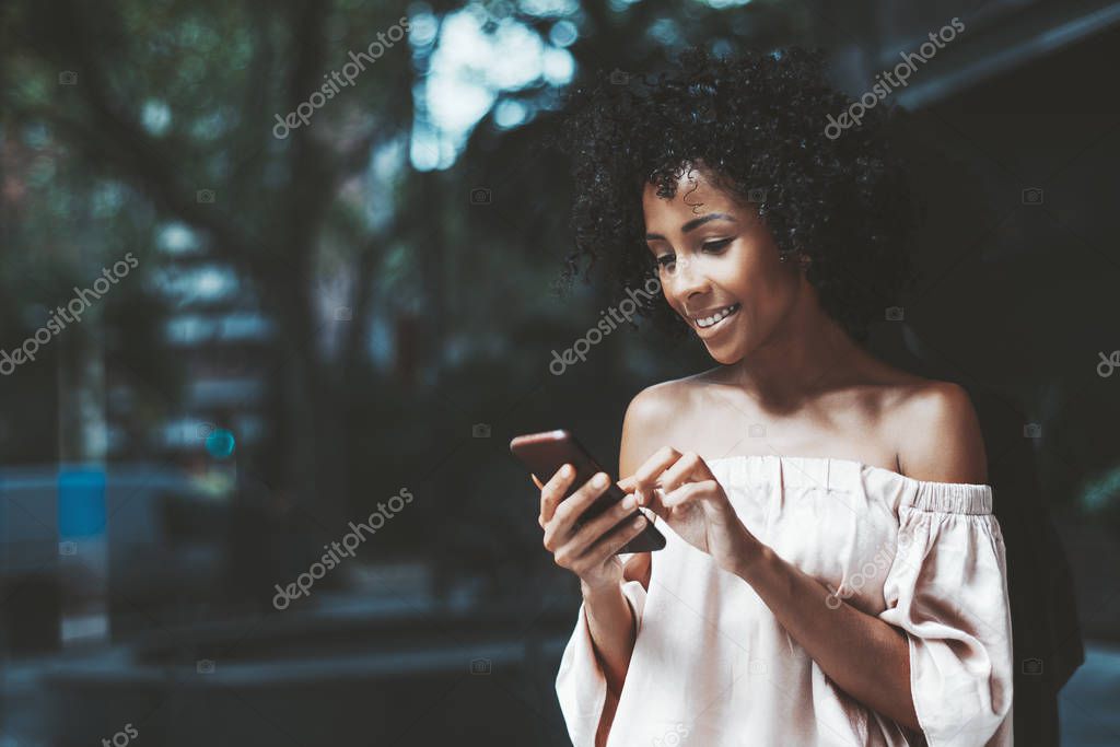 A happy young biracial female is standing next to a glass wall on a street and typing a message to her friend using the smartphone, with a copy space place on the left for your logo or a message