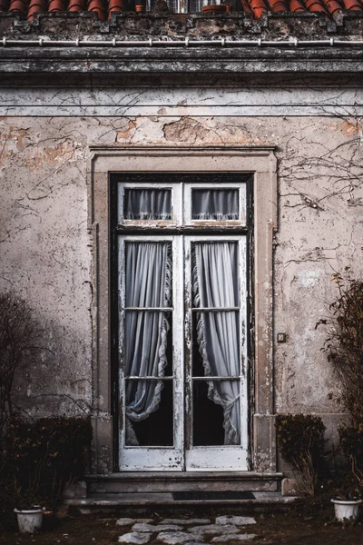Vertical shot of an antique porch with a peeling wooden paint frame and old white lace curtains inside, surrounded by the wall with partly flaked plaster; the typical architecture of Sintra, Portugal