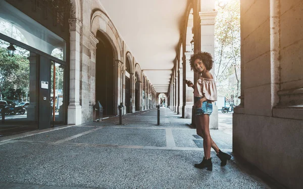 美しい若い毛アフリカ系アメリカ人女性立っているアーチとアンティークなコピー スペースと 建物の柱の屋外ホールでスマート フォンでの広角ショット — ストック写真