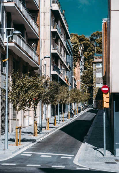 A short narrow one-way road in a residential district of Barcelona with a stop sign on the traffic light in front, the row of neatly planted trees, dwelling houses around, the hill in the distance