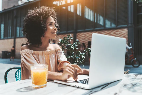 Düşünceli bir göz kamaştırıcı genç Brezilyalı kadın ikindilerde bir mesafede bir şey ise onu laptop ile sokak bir barda oturan ve serbest bir proje, bir bardak meyve suyu yanında üzerinde bakıyor