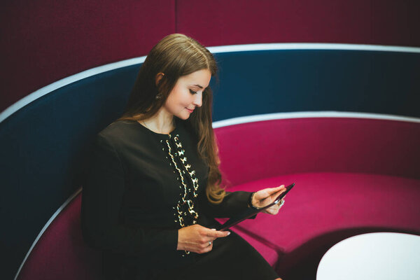 A charming young hispanic woman entrepreneur is sitting on a lilac round sofa and reading analytics on the screen of a digital tablet in her hands, with a copy space place on the right for an ad text