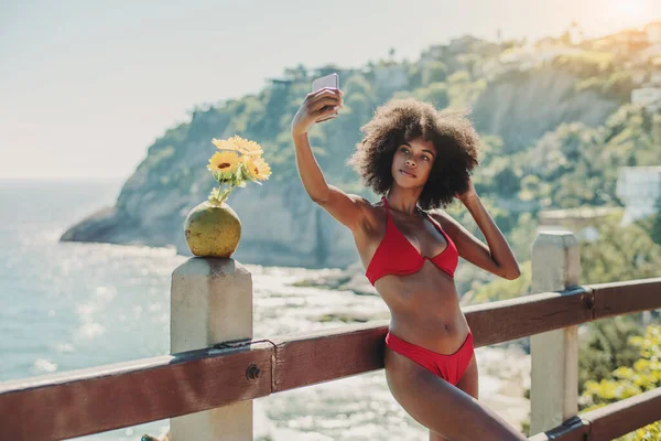 ビーチの近くのリゾートの木製の柵にもたれている間 セクシーな若いブラジル人女性はスマートフォンを使って自撮りしています ココナッツと彼女の近くの花 アフリカ系アメリカ人の女の子写真を撮る — ストック写真