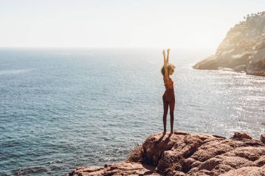 Kıvırcık saçlı göz kamaştırıcı genç Afro-Amerikalı kadın, kıyı şeridi kayasında dururken güneşin tadını çıkarıyor. Yazlık bir tatil köyünün dağında kollarını kaldırmış çekici Brezilyalı kız.