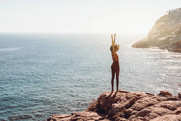 Kıvırcık saçlı göz kamaştırıcı genç Afro-Amerikalı kadın, kıyı şeridi kayasında dururken güneşin tadını çıkarıyor. Yazlık bir tatil köyünün dağında kollarını kaldırmış çekici Brezilyalı kız.