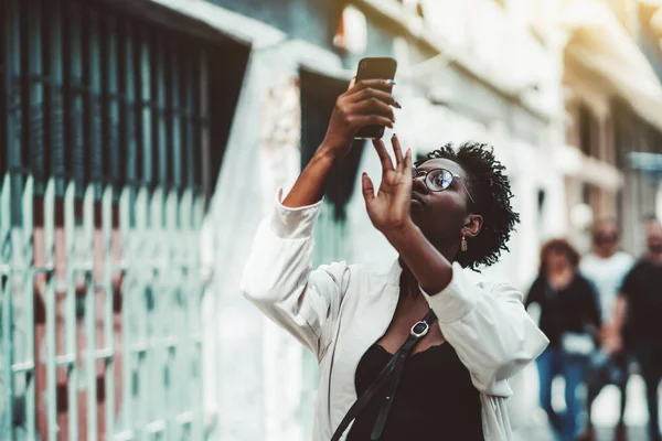 身穿眼镜 头罩和白色战壕的年轻时尚的非洲女性游客正在一条生机勃勃的街上用智能手机拍照观光 而黑人女孩则用手机拍照 — 图库照片