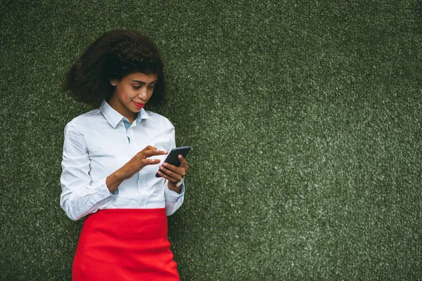 A charming African-American woman with a cellphone is standing in front of a green wall of artificial grass; young biracial businesswoman is using her smartphone in front of the wall of plastic grass