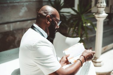 Siyah, düzgün sakallı, gözlüklü, beyaz takım elbiseli, yaz güneşiyle aydınlanan ve birden fazla yer imi olan bir kitap okuyan dalgın, kel ve olgun bir Afrikalı adamın yan görüntüsü.