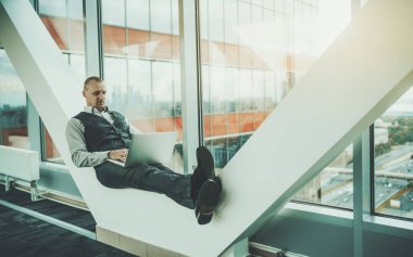 Kendine güvenen Kafkasyalı bir iş adamı, bir bankta otururken dizüstü bilgisayarı üzerinde çalışıyor, modern ofis binasının destekleri ile pencerenin önündeki bina cephesini birleştiriyor.