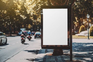 Yolun yakınındaki bir kaldırım taşında boş beyaz reklam panosu şablonu; kaldırımda boş reklam afişi taşıyıcı şablonu; sokak aralığının gölgesinde kentsel ayarlarda poster modeli