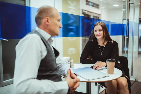 Business conversation of two colleagues inside of a coffee point and kitchen of a modern office: a businesswoman listening to her partner who sitting in front of her with a laptop and a disposable cup