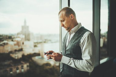 Bir iş yeri binasının panoramik penceresinin yanında duran ve akıllı telefonunu kullanan ciddi odaklı bir girişimcinin yan görüntüsü.