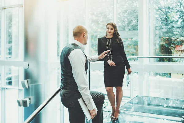Two entrepreneurs having a conversation on the top of a modern glass office stairway: a man with a laptop in his hand is telling his female partner with a digital tablet in hand news about a business
