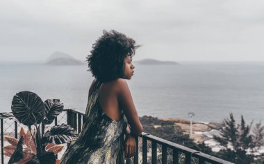 Göz kamaştırıcı genç, uykulu Brezilyalı bir kız balkon çitlerine yaslanıyor ve Rio de Janeiro 'nun yağmurlu bir manzarasına bakıyor. Arpoador plajı ve sönmüş arka planda bulutlu bir gökyüzü.