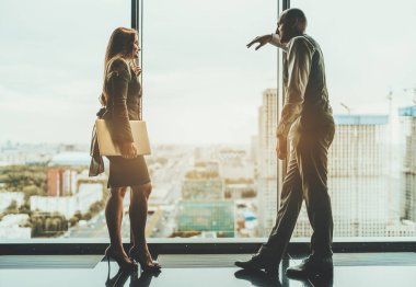 İnşaat alanı ve panoramik pencerenin dışında otoyolu olan şehir silueti olan iki iş arkadaşı silueti: panoramik pencerenin dışında bir erkek girişimci dizüstü bilgisayarlı kadın ortağıyla konuşuyor.