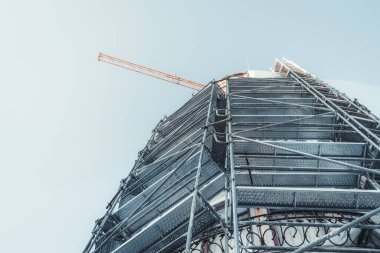 Yapılandırma işlemi sırasında binanın altından tüm cephesine monte edilmiş birden fazla metal iskeleye ve arkasında mavi bir gökyüzü olan bir inşaat vincinin bir kısmına bakın.