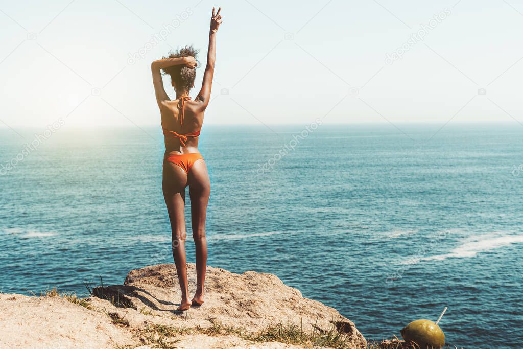 View from behind of a svelte sexy African-American female on a coastline cliff, she is stretching up, while standing up on her toes and showing a victory sign with her fingers on; ocean horizon behind