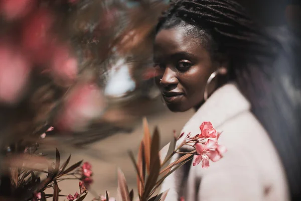 一个迷人的年轻黑人女人站在公园里 用辫子看着镜头 有选择性地将焦点集中在她的脸部和她面前的粉色花朵上 这是一张真实的倾斜变换画像 — 图库照片