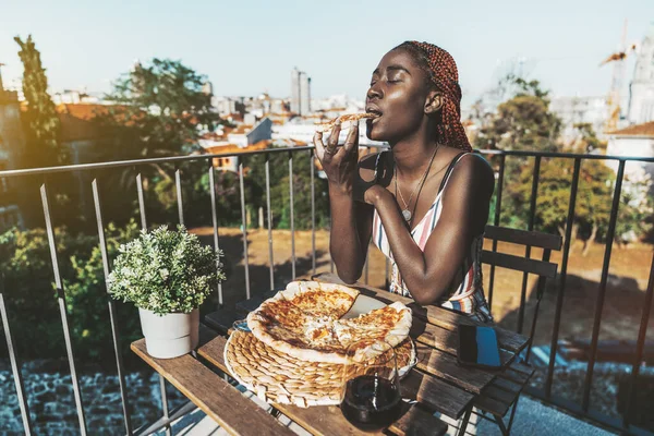 An adorable black girl eating a piece of a delicious vegan pizza while sitting on a balcony of a street pizzeria on the second floor with a glass of wine and a smartphone next to her, sunny morning