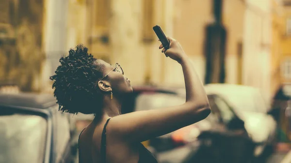 一个年轻的黑人女性用她的智能手机在街上拍照的侧影 复古色彩 戴着眼镜的迷人的非洲旅游女孩正在用她的手机拍摄外面的东西 — 图库照片