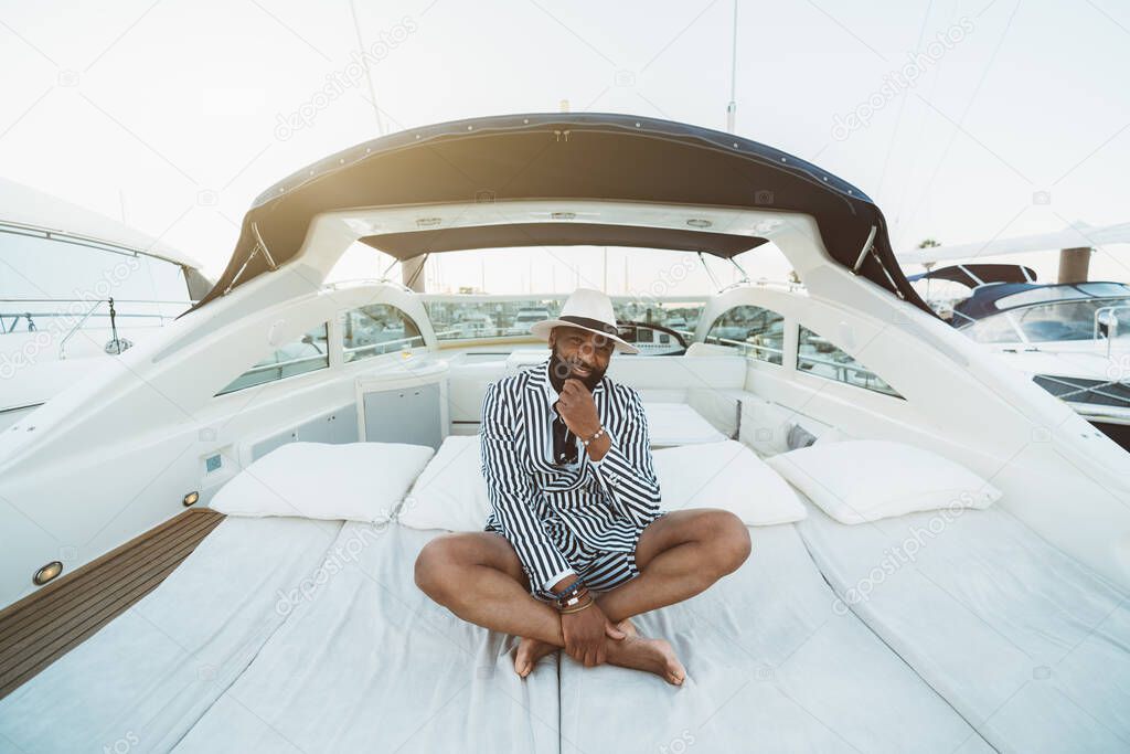 A cheerful wealthy bearded mature black guy in an elegant striped black and white costume with shorts and hat is sitting outdoors on the deck of a luxury bright speed yacht moored on a berth