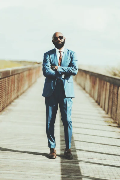Vertical shot of a handsome mature bald black man entrepreneur in a formal elegant custom made blue suit, sunglasses, necktie; he is standing in the middle of a long wooden bridge with hands crossed