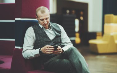 Yakışıklı bir İspanyol girişimci, modern bir ofis ortak çalışma alanının leylak kanepesinde otururken, akıllı telefonuyla sohbet konuşmalarını okurken gülümsüyor; sağ tarafta bir fotokopi yeri.