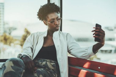 Güneş gözlüklü, kamuflaj pantolonlu, beyaz siperli, genç sakin bir kadının portresi. Halat kulübesinde bir bankta oturuyor ve akıllı telefon kamerasıyla selfie çekiyor.
