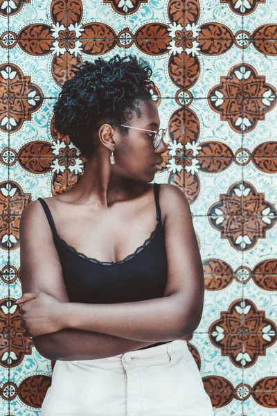 一张垂直的照片 是一个年轻可爱的黑人女性 身穿水罐上衣和眼镜 一头卷曲的非洲头发 站在葡萄牙传统的阿苏莱霍瓷墙前 望着旁边 — 图库照片