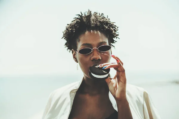 Portrait of a young beautiful black woman in sunglasses and a white trench, holding in her hand with nail-art and eating delicious ice cream inside of two black biscuits while standing outdoors