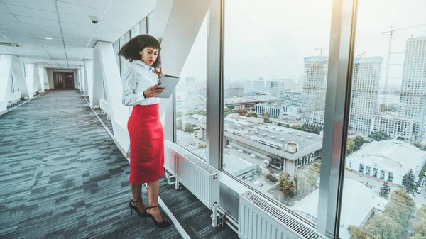 一位身穿白衬衫和红色裙子的美丽的非洲裔美国女企业家站在办公室走廊的窗前 一边在她的数码平板电脑屏幕上看工作聊天 — 图库照片