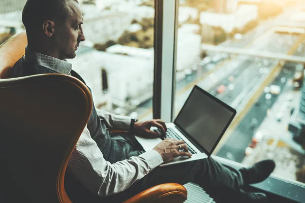 Side view of a mature handsome caucasian man entrepreneur working on his netbook while sitting in a comfortable luxury office area on an orange armchair near the window of a modern business skyscraper