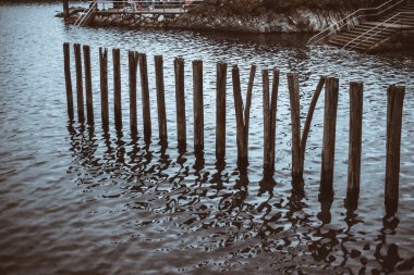 Taş iskele ve merdiven kenarındaki sakin dalgalı sularda duran eski ahşap direkler yansımalar ve kırsal bir deniz atmosferi yaratıyor.