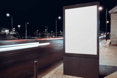 Blank vertical billboard mockup on city street at night with glowing lamps, light trails from passing traffic and modern urban background, perfect for advertising design showcase; long exposure
