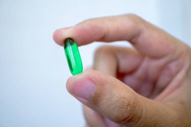 Tek bir yeşil oval kapsülü tutan bir insan elinin yakından görüntüsü, muhtemelen bir beslenme takviyesi ya da ilaç. Arka plan sade, kapsüle ve elin hareketine odaklanmış durumda.