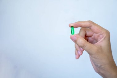Tek bir yeşil oval kapsülü tutan bir insan elinin yakından görüntüsü, muhtemelen bir beslenme takviyesi ya da ilaç. Arka plan sade, kapsüle ve elin hareketine odaklanmış durumda.