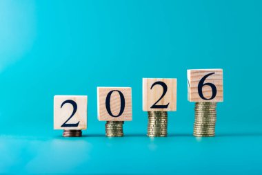 2026 numaralı tahta bloklar turkuaz mavi arka plan üzerine yerleştirilmiş bozuk para yığınları, yatay kompozisyon. Uzayı kopyala Finans, tasarruf, yatırım ve iş büyümesi kavramı