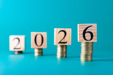 Yığılmış paralara göre 2026 numaralı tahta bloklar oluşturuluyor, 6 numaraya odaklan, turkuaz arka planda yatay görüntü. Tasarruf, para büyümesi ve yeni fırsatlar kavramı