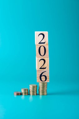 2026 'yı oluşturan tahta blokların dikey çekimi mavi arka planda para yığınları ile. Uzayı kopyala Tasarruf, finans, ekonomik büyüme ve yeni yıl kavramı