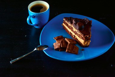 Mavi tabakta bir parça çikolatalı pasta. Bir fincan kahvenin yanında. Hepsi siyah ahşap bir arka planda..