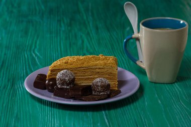Bir parça kek ve bir tabakta çikolata, yanında bir fincan kahve, yeşil ahşap bir arka planda yakın plan fotoğrafı..