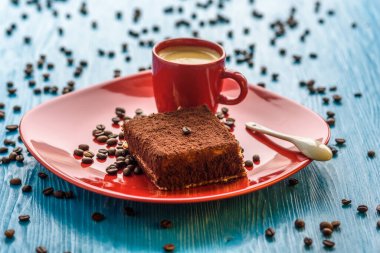 Kırmızı bir tabakta tiramisu keki, bir fincan kahvenin yanında, açık ahşap bir arka planda fotoğraflanmış..