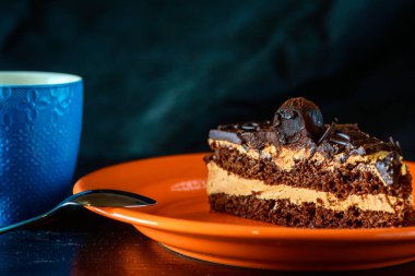 Bir parça çikolatalı kek turuncu tabakta, mavi bardaktaki kahvenin yanında..