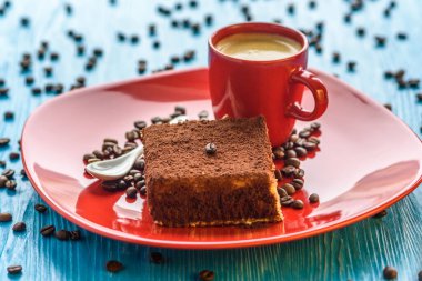 Kırmızı bir tabakta tiramisu keki, bir fincan kahvenin yanında, açık ahşap bir arka planda fotoğraflanmış..
