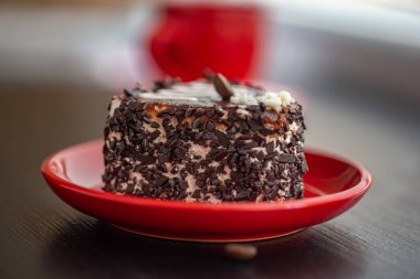 Kırmızı bir tabakta çikolata parçaları serpiştirilmiş kalp şeklinde lezzetli bir pasta. Ahşap bir arka planda yakın plan çekilmiş..