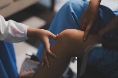 Kadın bir doktor, eklem iltihabı nedeniyle diz ağrısı olan yaşlı bir hastayı muayene eder. Hastaya iyileşme sürecinde yardım ediliyor, ağrı yönetimi ve hareket kabiliyetinin iyileştirilmesi üzerinde duruluyor..