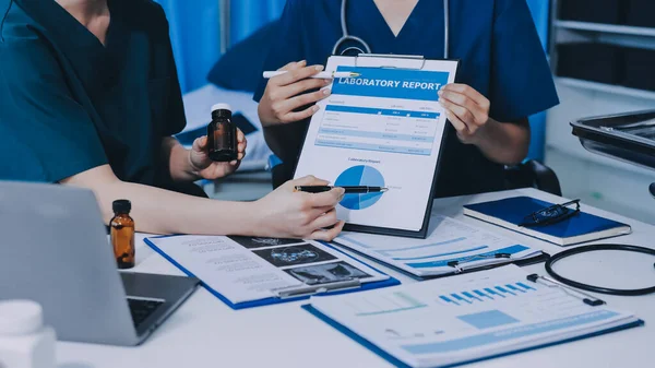 Two doctors and a female nurse meet at a table in the hospital, collaborating on medical tasks using laptops and computers