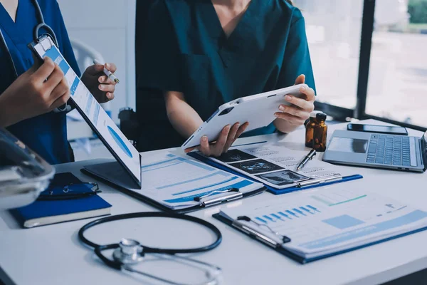Two doctors and a female nurse meet at a table in the hospital, collaborating on medical tasks using laptops and computers