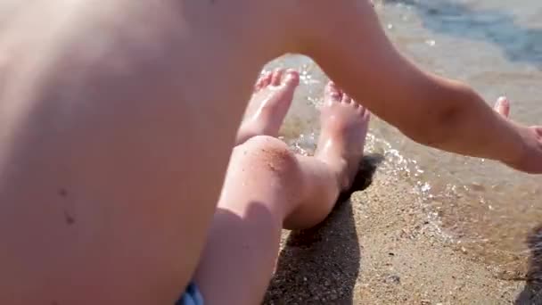 Un garçon est assis au bord de la mer et attrape les vagues. Les vagues lavent les pieds des garçons. Vacances en mer pour la famille avec enfants . — Video